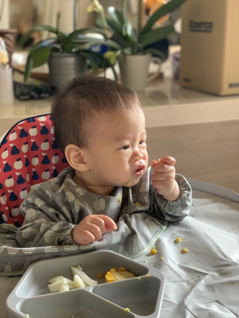 A baby boy wears Tidy Tot bib, eating baby food on a baby feeding chair