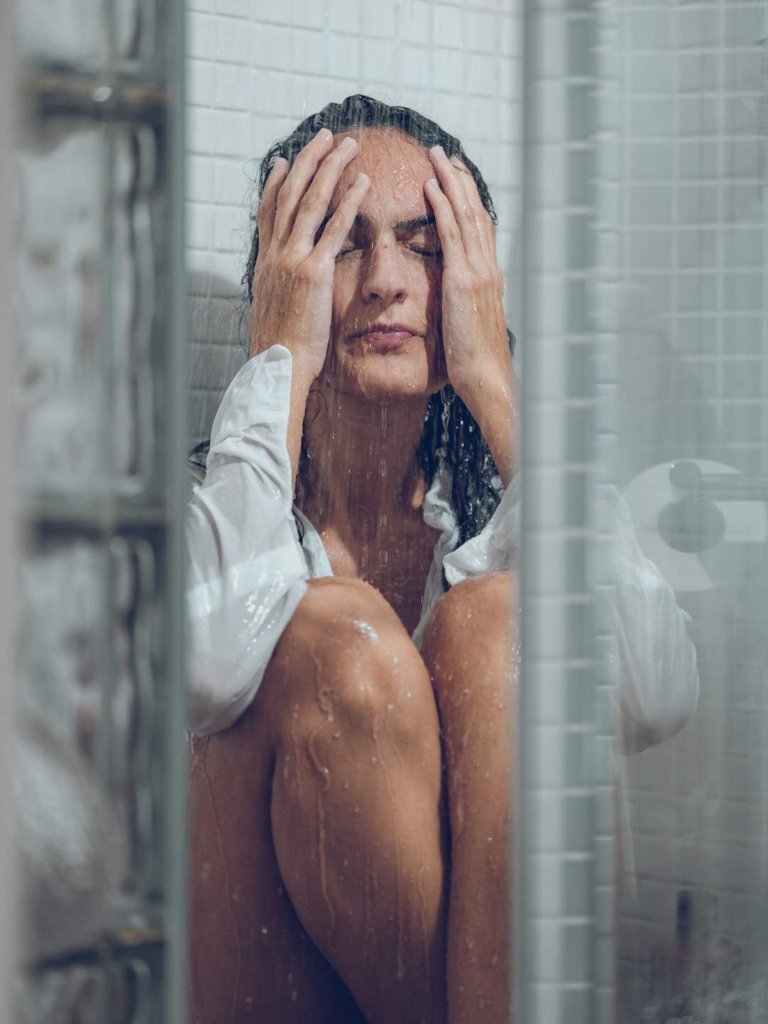 Sad female with wet dark hair wearing white shirt sitting in tiled shower cabin under water stream with closed eyes and touching head