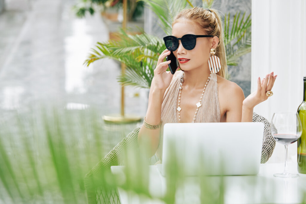 Rich gorgeous young woman in sunglasses talking on phone when working on laptop in outdoor cafe